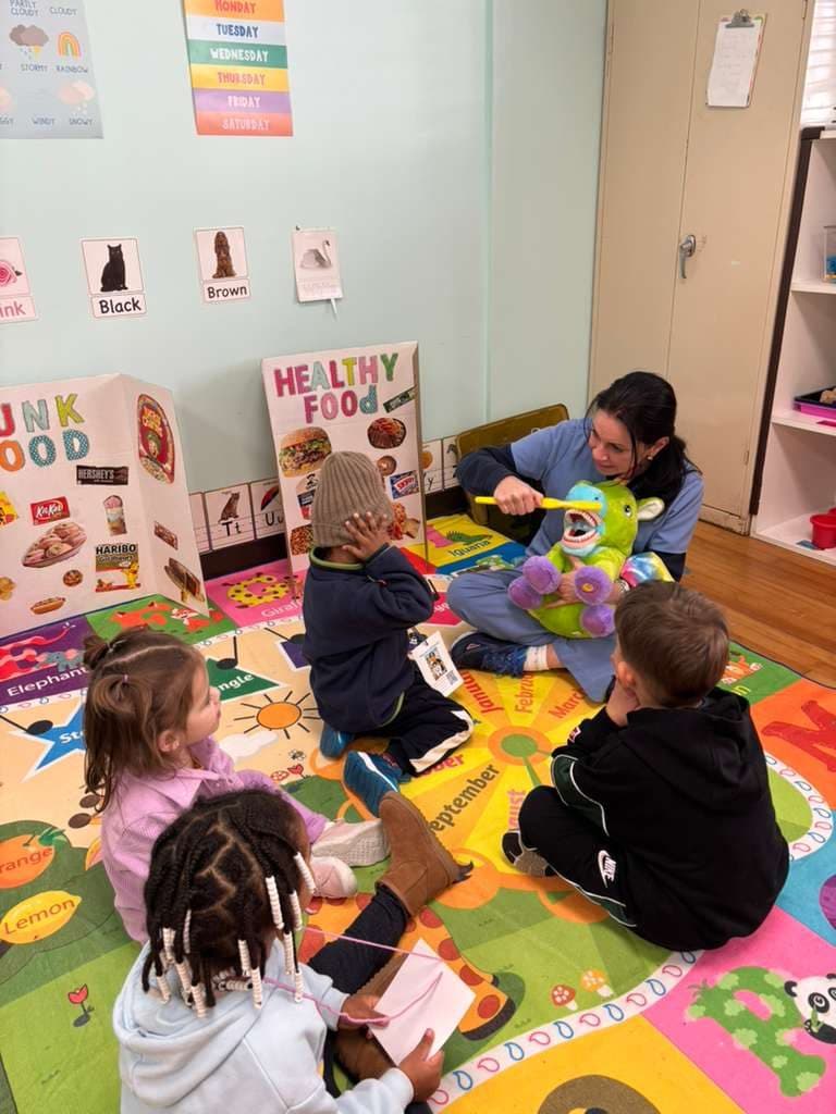 A teacher shows a group of children how to brush teeth, using an oversized toothbrush and a soft toy.