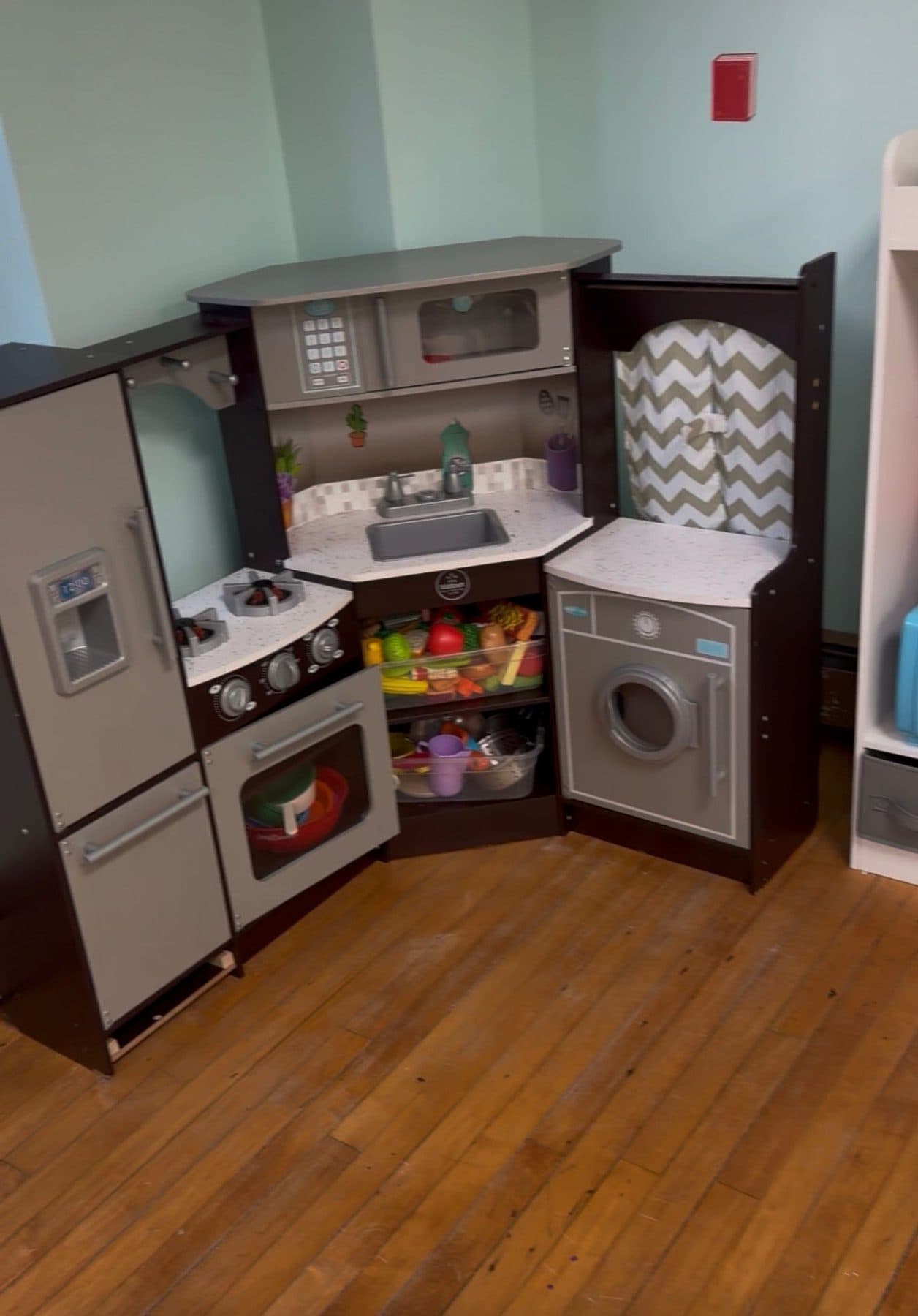 A wooden play kitchen stocked with toy food, pans, and a play washing machine.