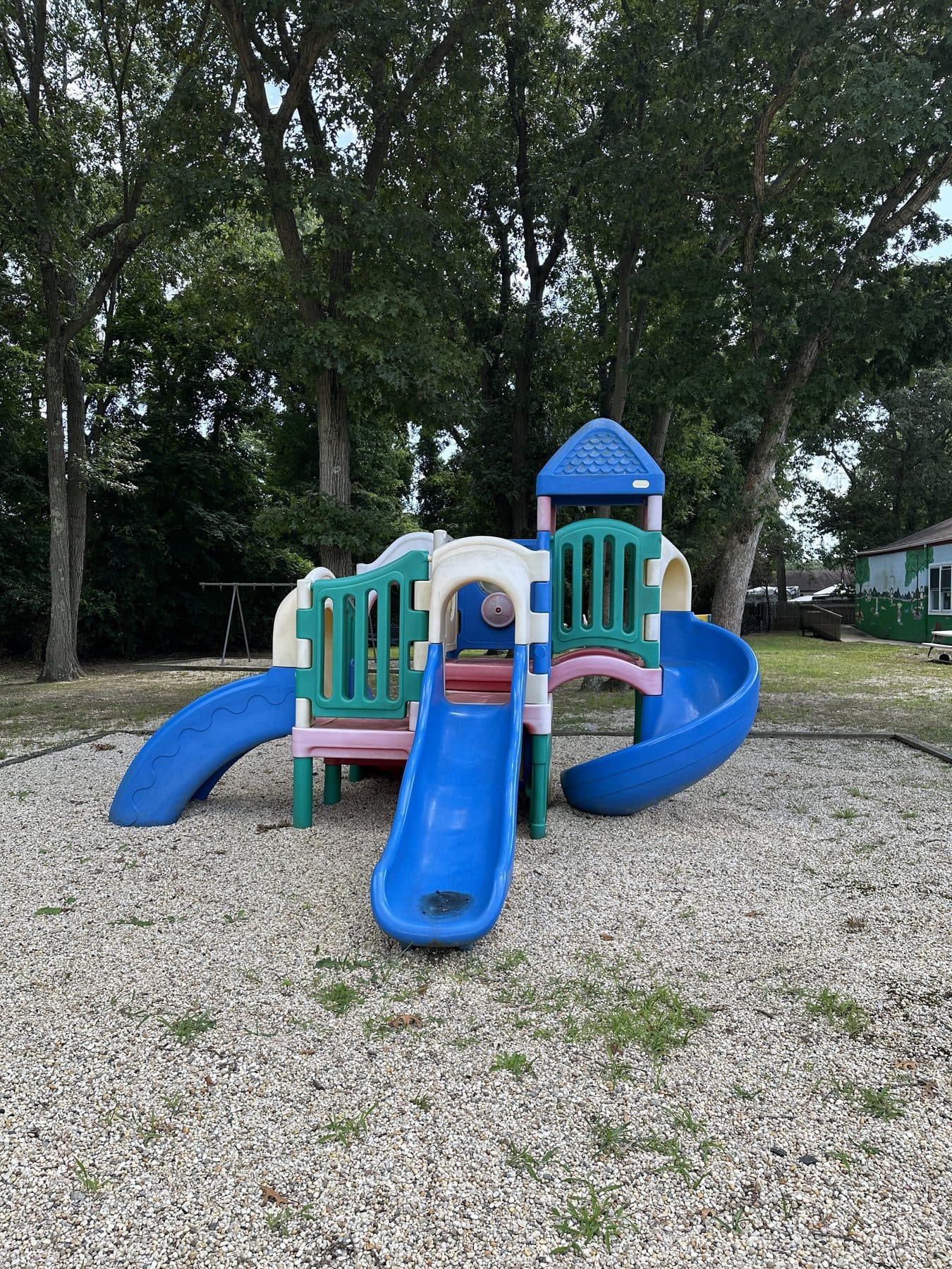 The outdoor playground: a climbing frame with two slides on gravel, surrounded by mature trees.