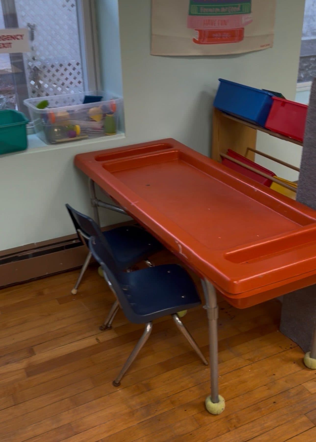 An orange sensory table set up by the window, with bins of materials on the sill behind it.
